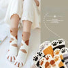 A pair of white plush socks with brown cat paw prints are shown on a person's feet, set against a soft background.