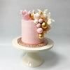 A pink buttercream cake decorated with gold and white floral toppers on a white pedestal stand.