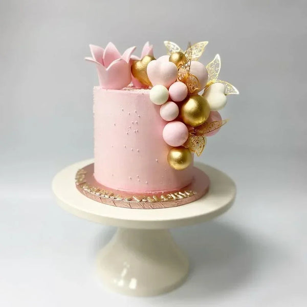 Pastel de buttercream rosa decorado con toppers florales dorados y blancos sobre un soporte blanco.