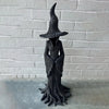 Estatua esbelta y oscura de una bruja con sombrero puntiagudo y túnicas fluidas, exhibida contra una pared de ladrillo blanco