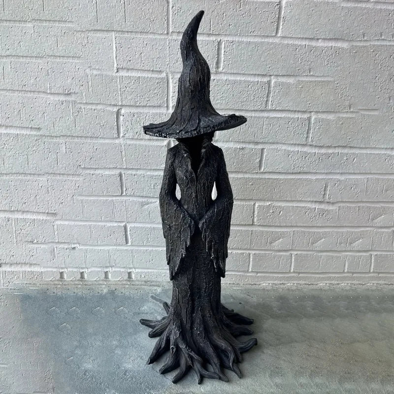 Estatua esbelta y oscura de una bruja con sombrero puntiagudo y túnicas fluidas, exhibida contra una pared de ladrillo blanco