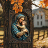 A metal sign depicting the Virgin Mary holding the infant Jesus, mounted on a tree trunk.