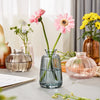 A modern clear glass vase filled with green stems and pink flowers sits on a light surface.