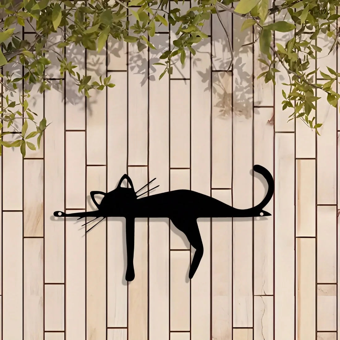 A black silhouette of a cat sculpture is mounted on a light wood panel with green foliage above.