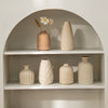 A display of minimalist ceramic vases and a dried flower in an arched shelf setting.
