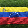 A large Venezuelan national flag banner featuring yellow, blue, and red stripes with stars and a coat of arms, displayed on g