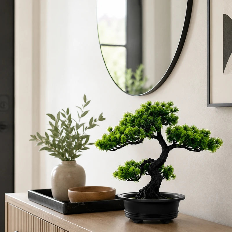 An artificial bonsai pine tree in a black pot sits on a wooden surface next to a mirror.
