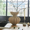 A beige ceramic vase with delicate branches sits on a wooden table near a window.