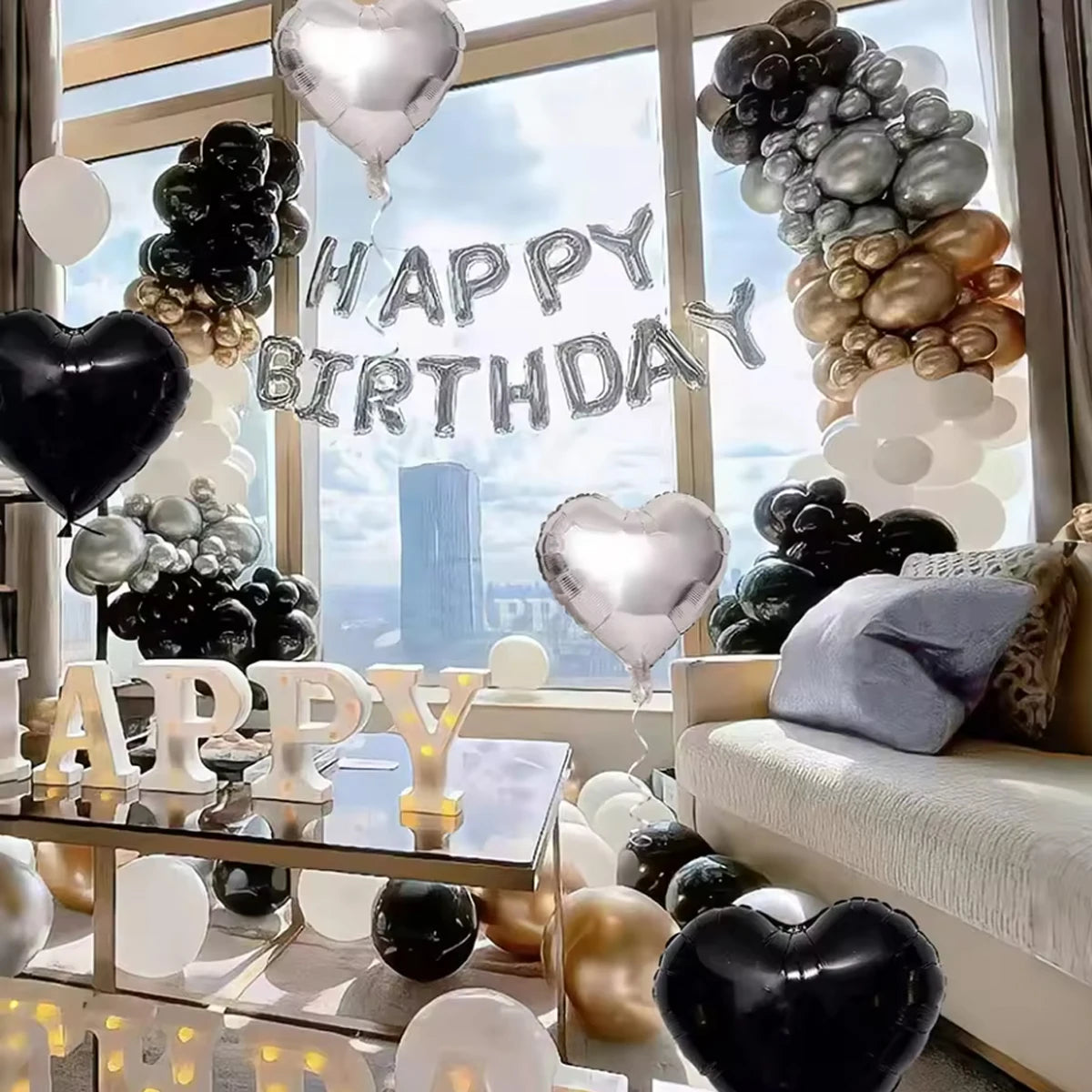 A display of black, white, and gold balloons arranged in clusters with a silver banner against a window backdrop.
