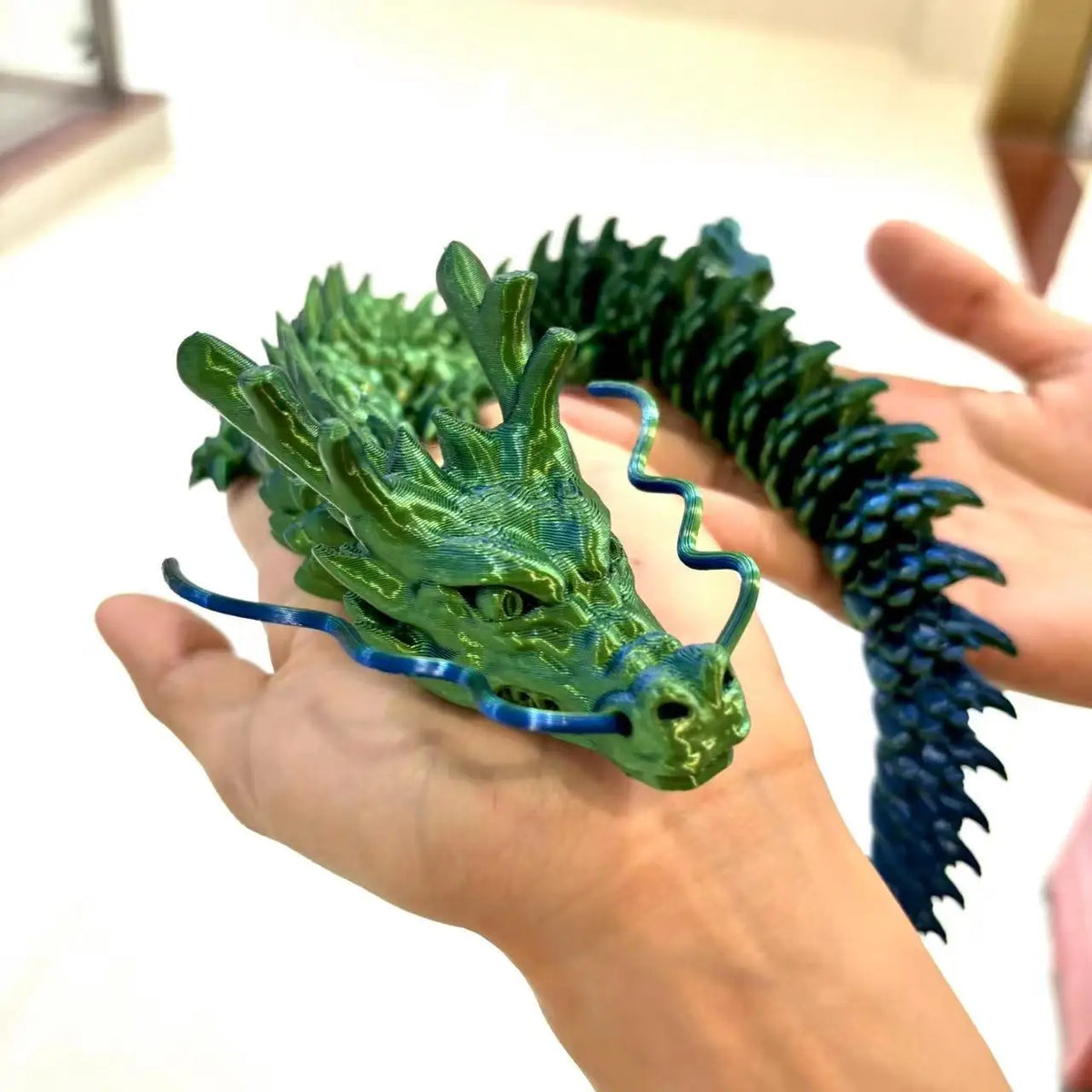 A hand holds a green articulated 3D printed dragon figurine with blue accents.
