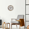 A wooden wall clock with a Roman numeral face hangs on a white wall next to a wooden chair and a small table.