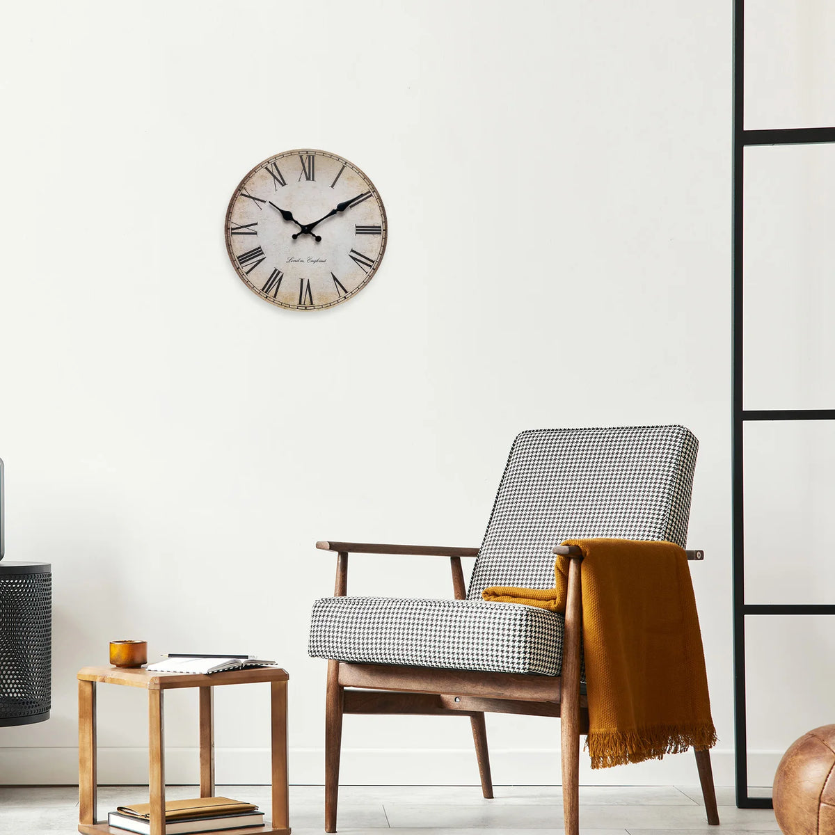 A wooden wall clock with a Roman numeral face hangs on a white wall next to a wooden chair and a small table.