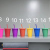 A row of fifteen brightly colored plastic cold cups with lids and straws displayed in a row.