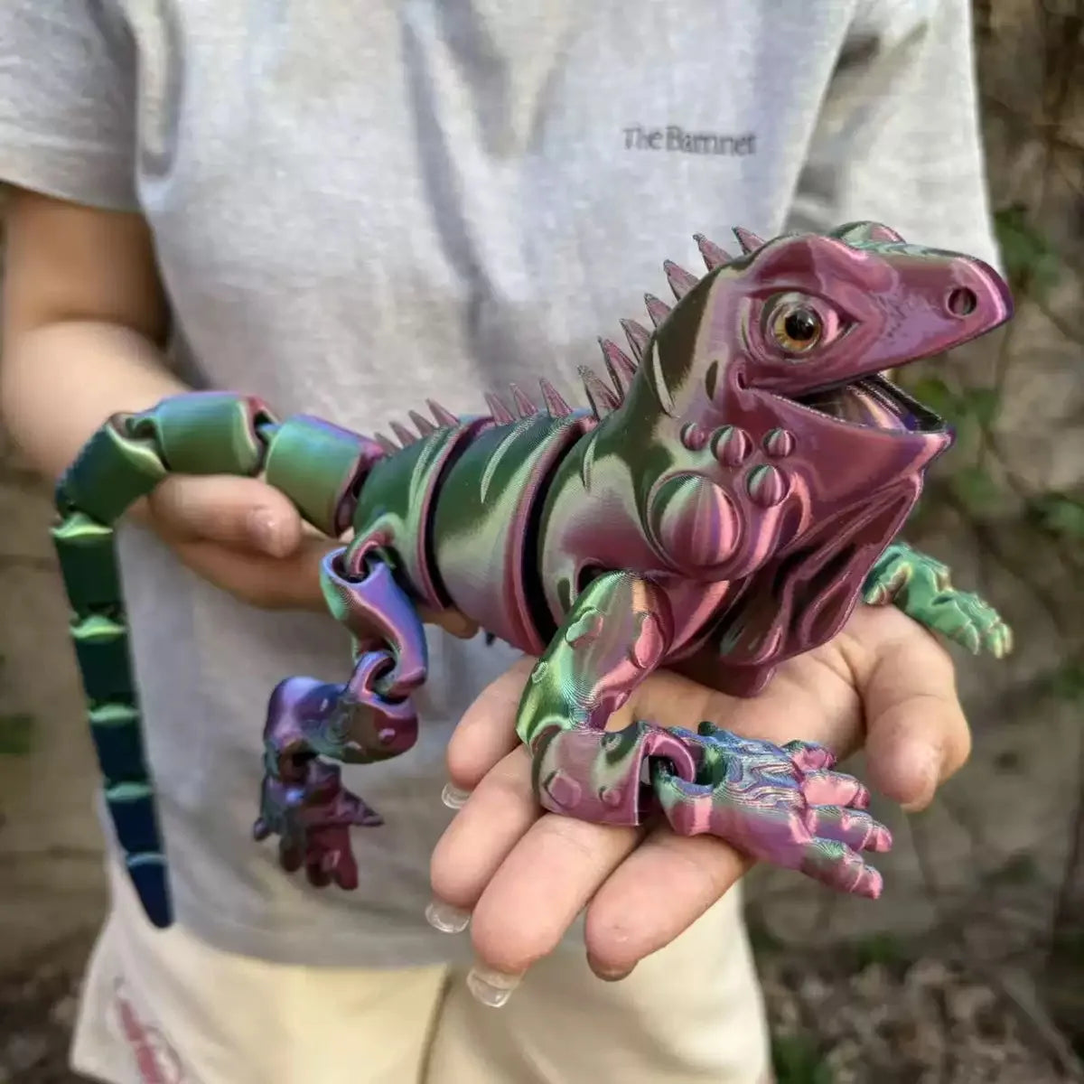 A hand holds a detailed, articulated 3D printed lizard figurine with purple and green scales.