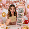 A young woman holds a stack of birthday cash gift envelopes in front of a pink cake and balloons.
