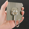 A hand holds a metal adhesive wall hook with two hooks, designed for home organization.