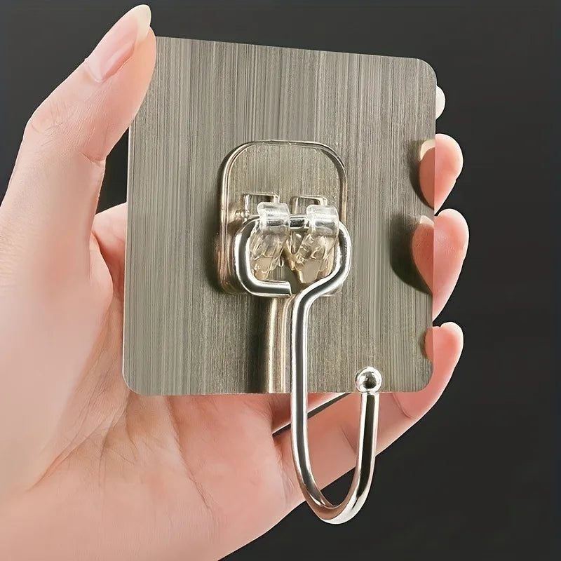 A hand holds a metal adhesive wall hook with two hooks, designed for home organization.