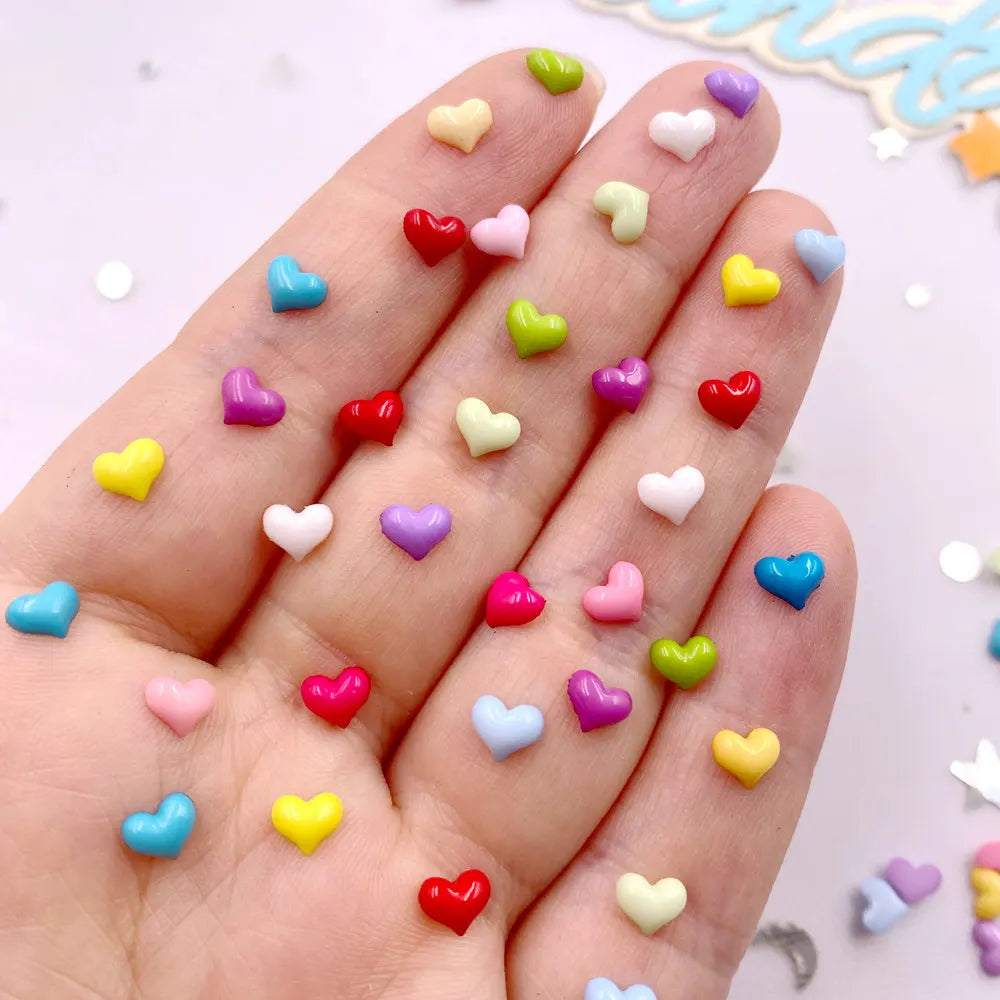 A hand displays a variety of small, flatback heart-shaped rhinestones in various colors for nail art and crafts.
