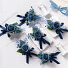 A set of six dusty blue silk rose boutonnieres with navy ribbon and white accents displayed on a white marble surface.