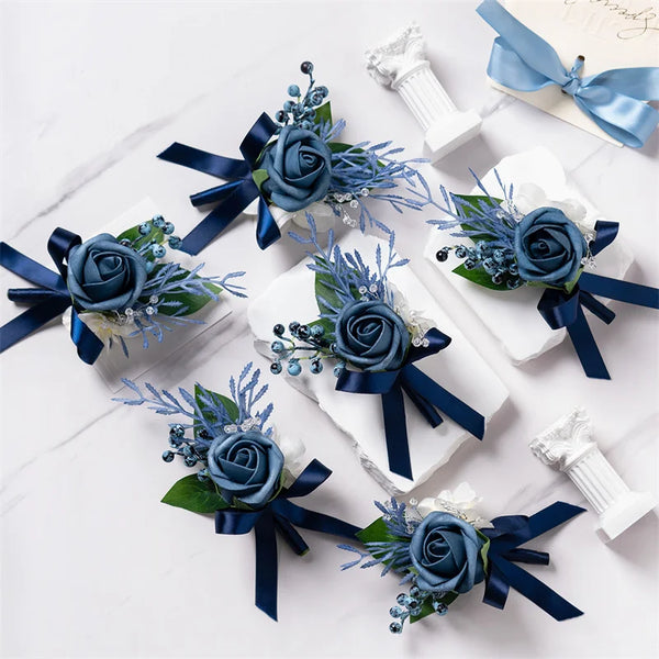 A set of six dusty blue silk rose boutonnieres with navy ribbon and white accents displayed on a white marble surface.