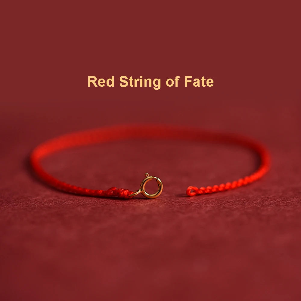 A close-up of a red string bracelet with a gold plated clasp lying on a dark red fabric background.