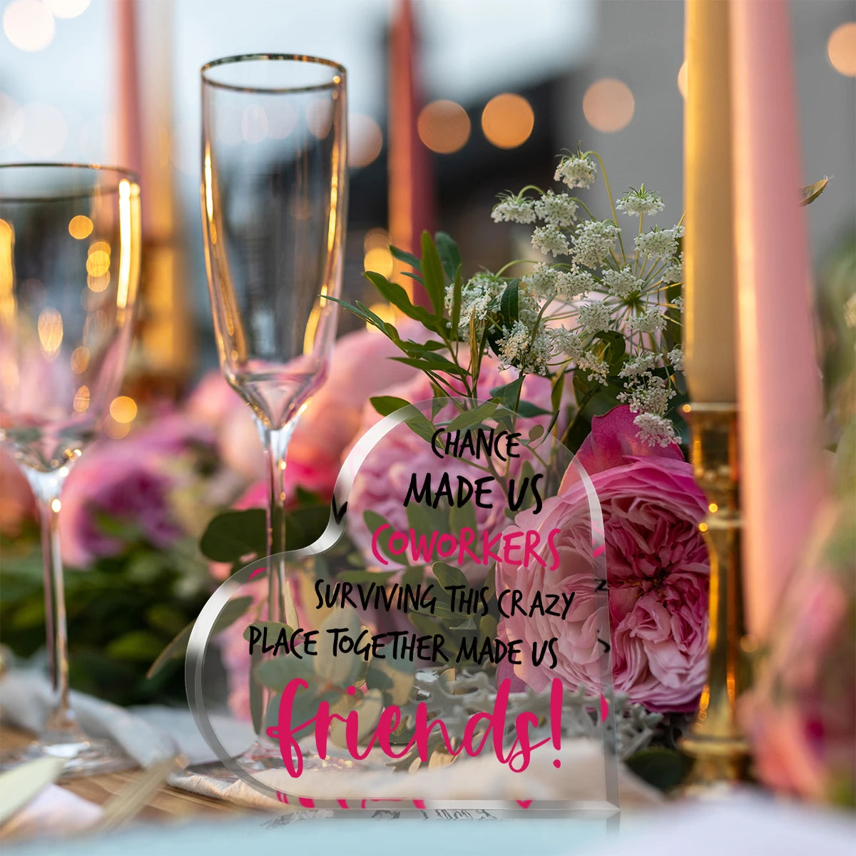 A heart-shaped glass plaque with pink lettering for a coworker friendship gift surrounded by flowers and candles.