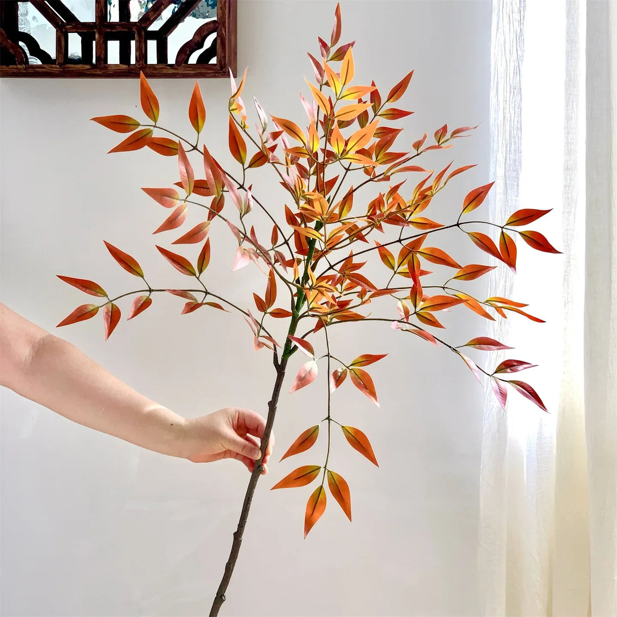 A hand holds a branch of artificial Nandina plant with reddish-orange leaves against a white background.