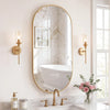 An oval wall mirror with a gold metal frame reflecting a bathroom interior and a sink area.