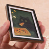 A framed print featuring a black cat looking at a slice of pizza against a dark green background.