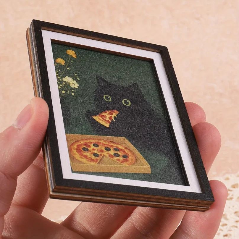 A framed print featuring a black cat looking at a slice of pizza against a dark green background.