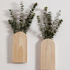 Jarrón de pared arqueado de madera con follaje verde, montado en una pared blanca.