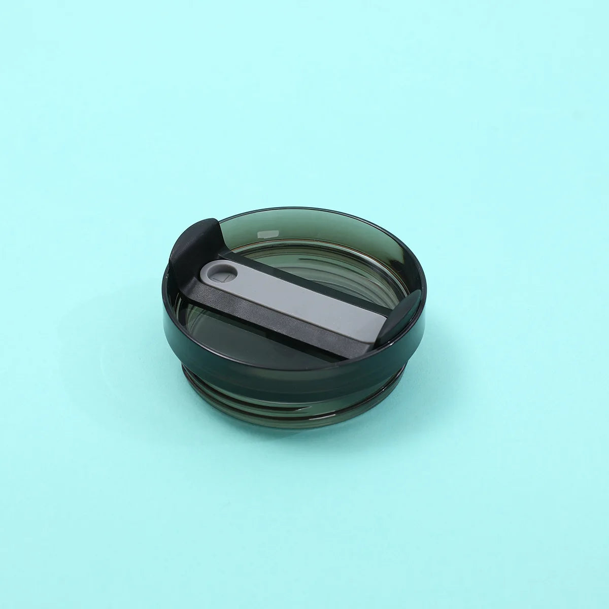 A dark green replacement tumbler lid with a silver-colored sliding mechanism resting on a light blue surface.