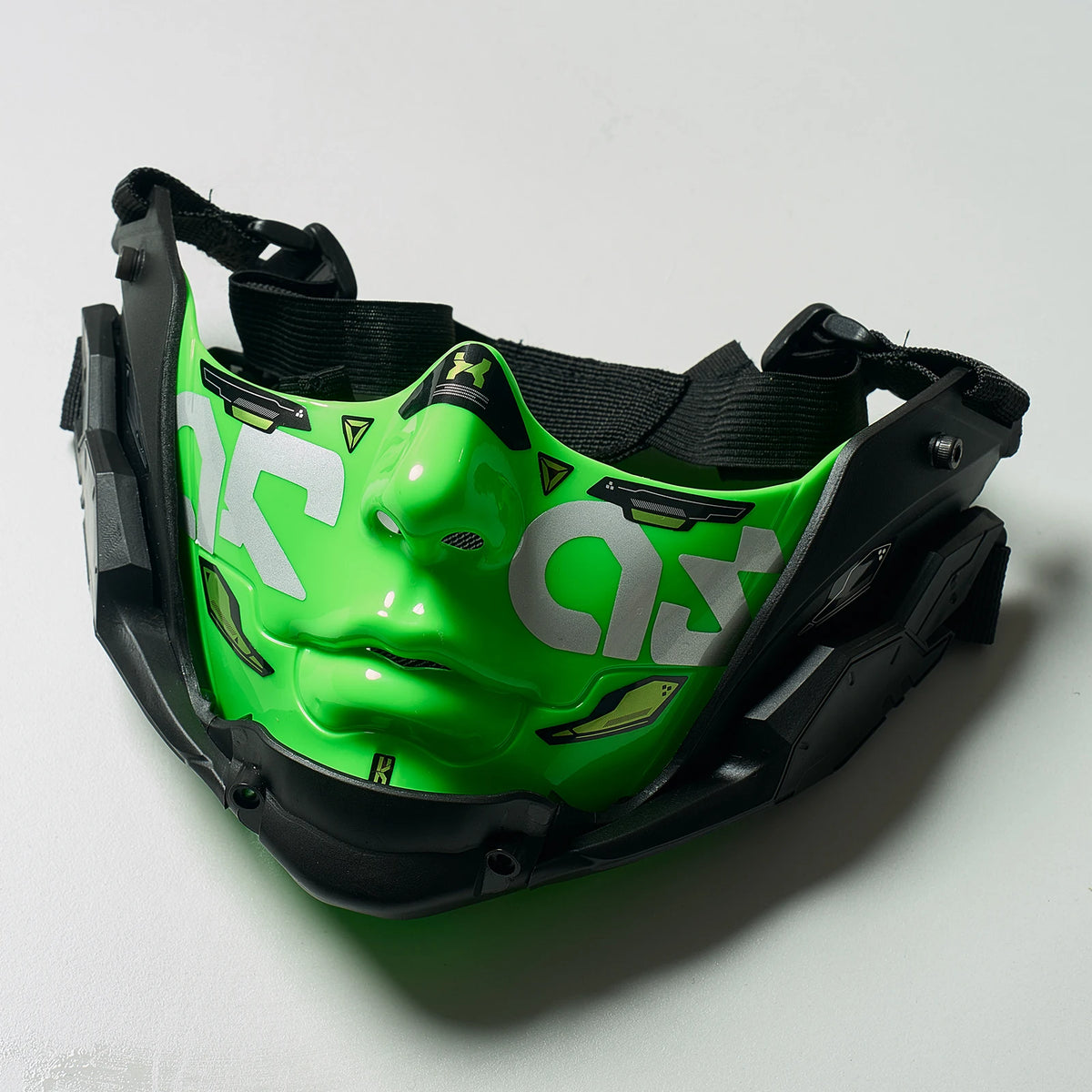 A green half face mask with a stylized eye design is shown in black protective gear.