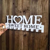 A hand holds a metal key holder with the words HOME SWEET HOME and wall-mounted hooks against a dark wooden background.