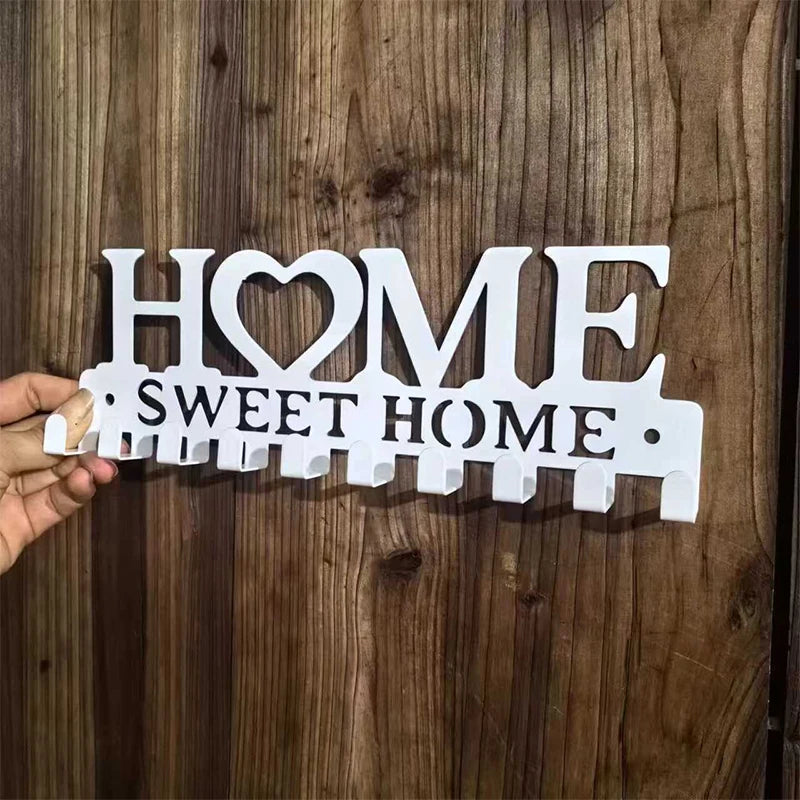 A hand holds a metal key holder with the words HOME SWEET HOME and wall-mounted hooks against a dark wooden background.