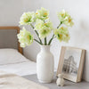 A vase holds several light green artificial clivia amaryllis flowers on a white surface next to a book.