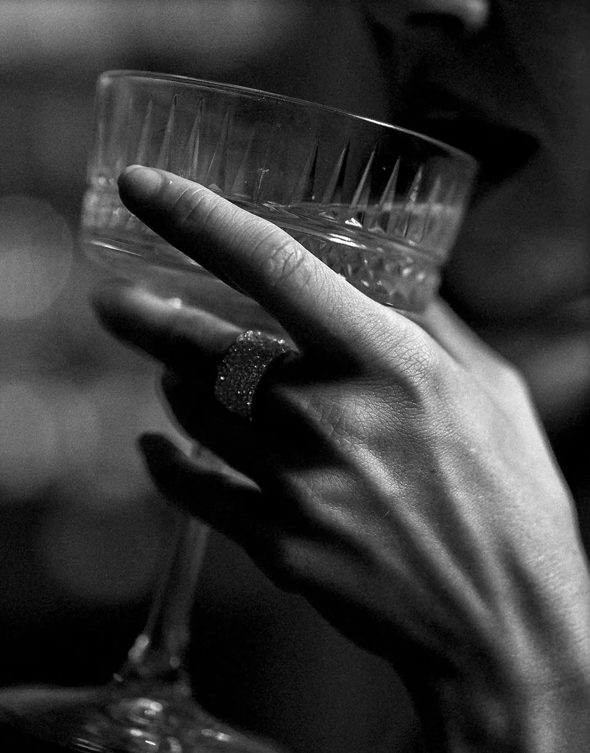 A hand holds a glass, featuring a ring, in a black and white photograph.