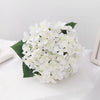 A bouquet of artificial white hydrangea flowers with green leaves on a white surface.