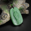 A green oval pendant depicting a figure, suspended from a dark beaded necklace and resting on a piece of textured wood.