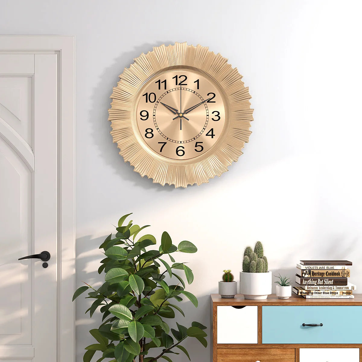 A round gold-toned wall clock with a sunburst design hangs on a white wall next to a wooden cabinet and a green plant.