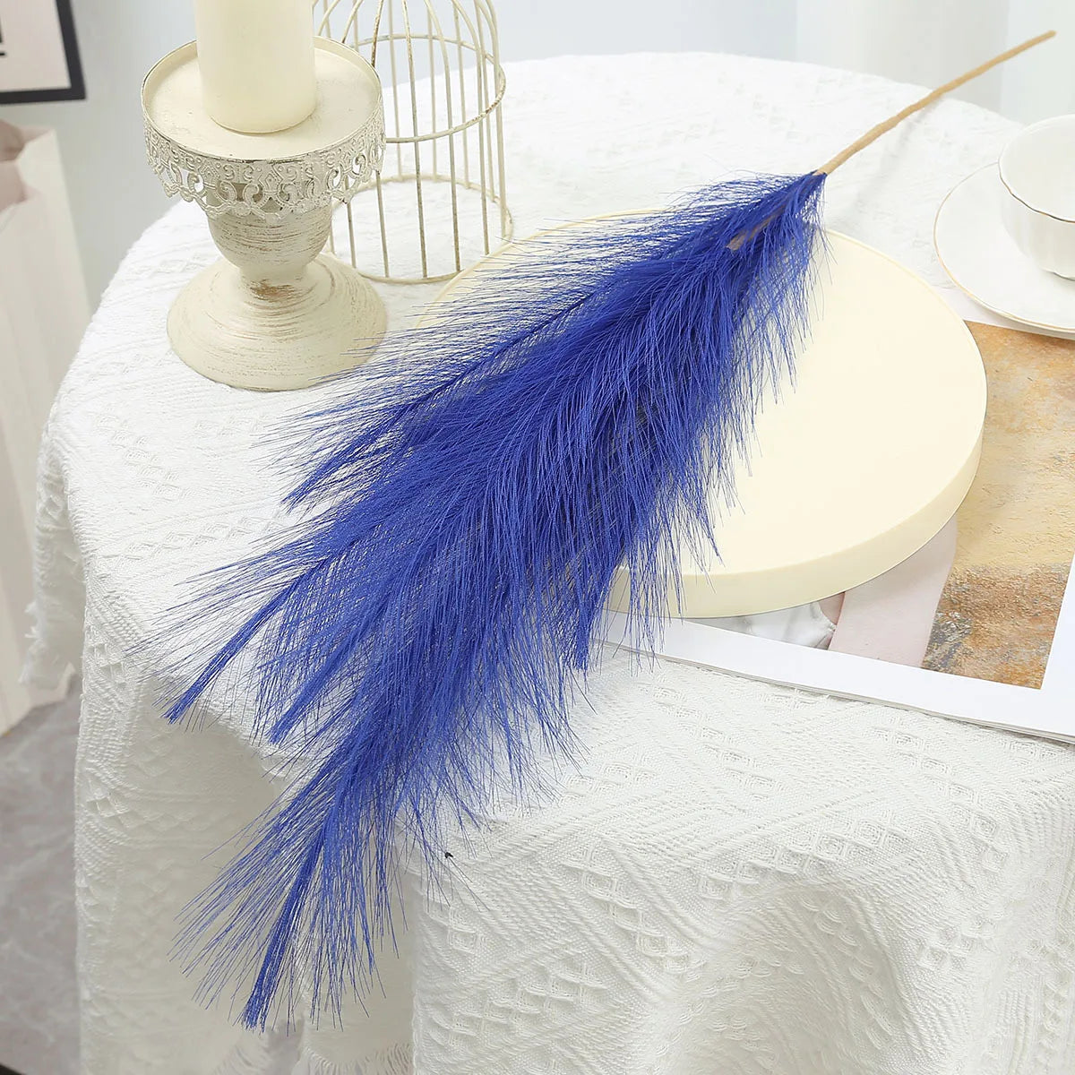 A vibrant blue artificial pampas grass feather displayed on a white linen tablecloth next to a decorative candle holder.