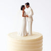 A bride and groom figurine topper made of white fondant standing on a tiered cake.
