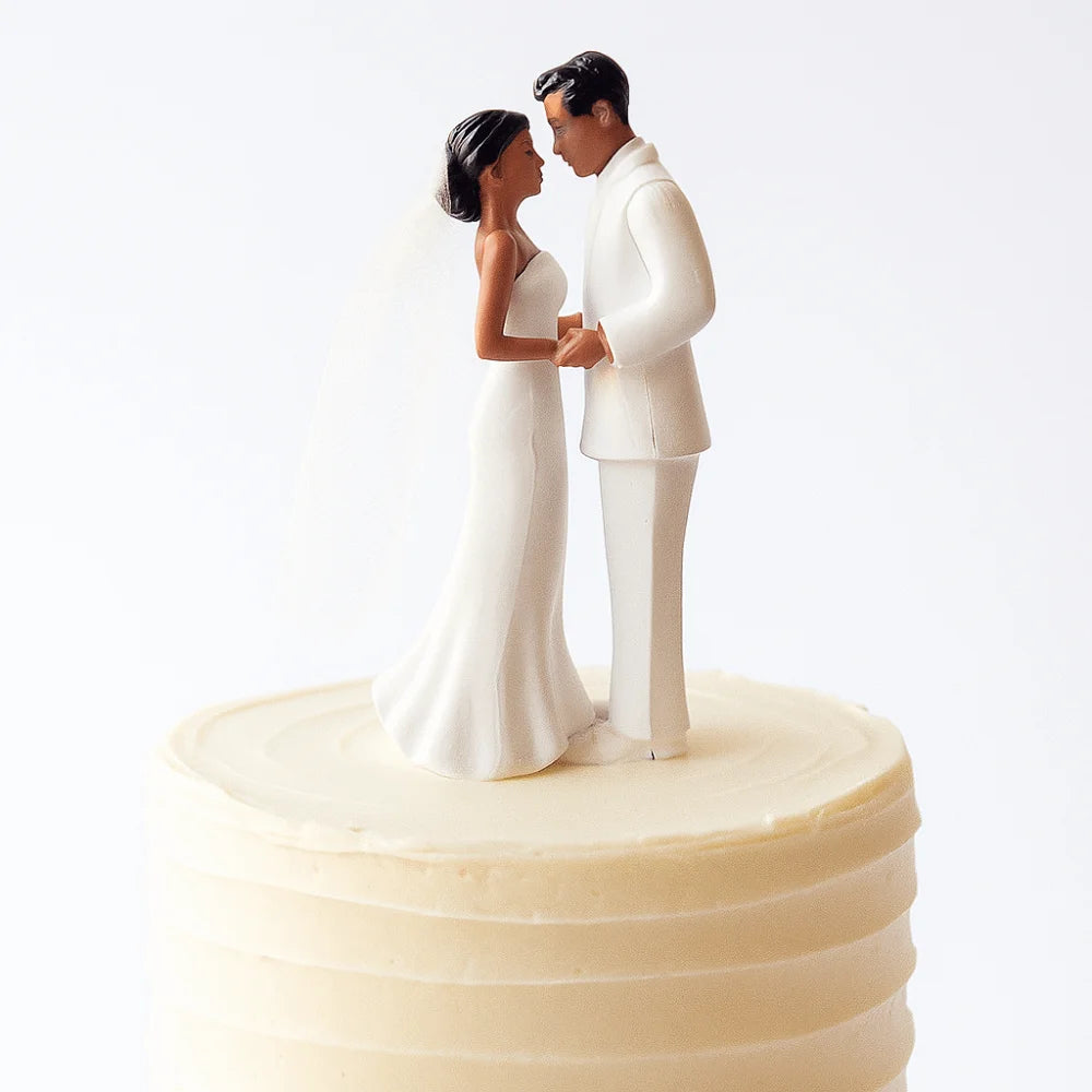 A bride and groom figurine topper made of white fondant standing on a tiered cake.