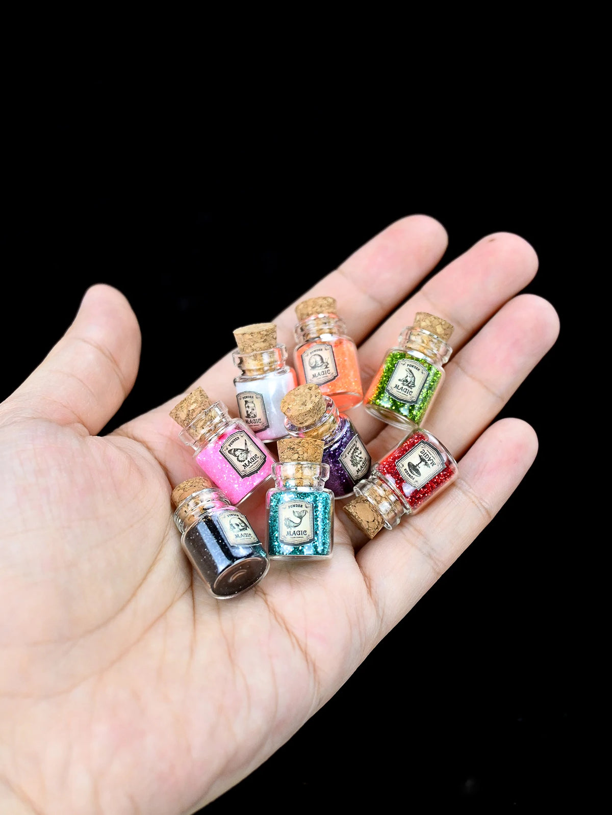 A hand holds several small glass bottles of glitter powder in various colors and textures for party decorations.
