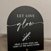 A black acrylic sign with white script that reads Let Love Glow and instructs to grab a glow stick and light up the dance flo