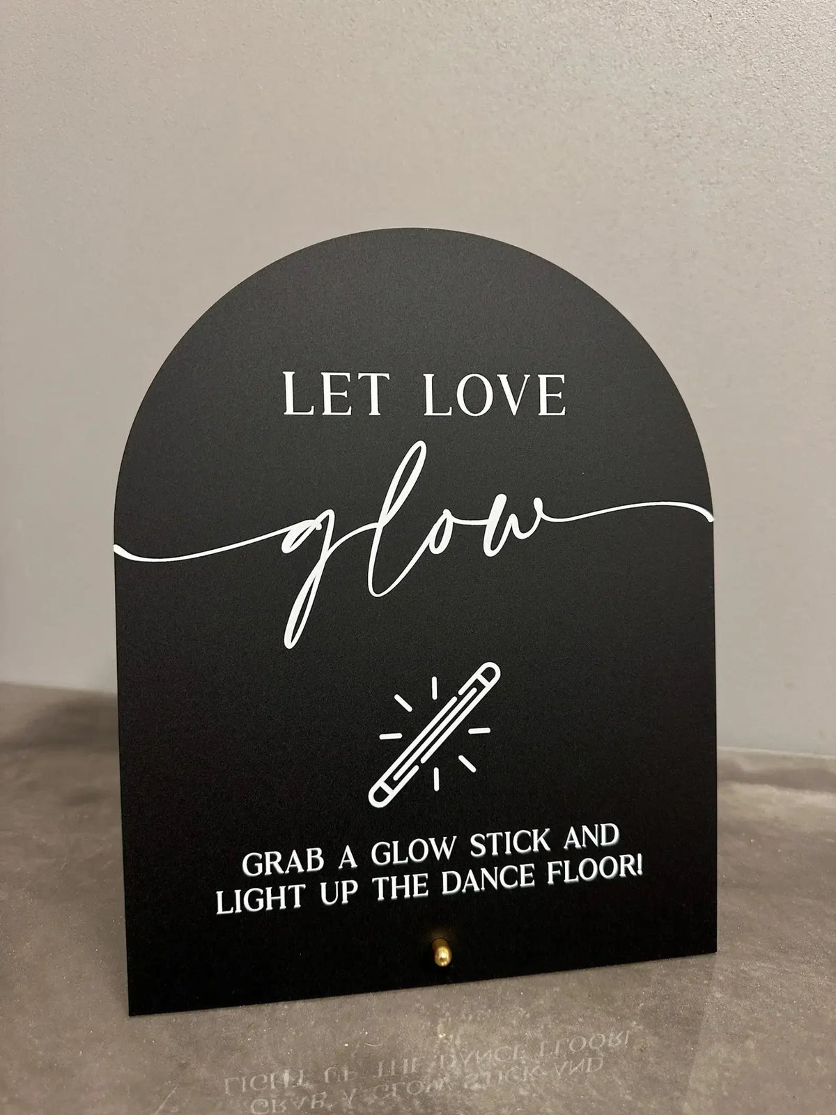 A black acrylic sign with white script that reads Let Love Glow and instructs to grab a glow stick and light up the dance flo