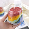 A hand holds a glass jar filled with layered colored sand pigment for a handmade candle wax kit.