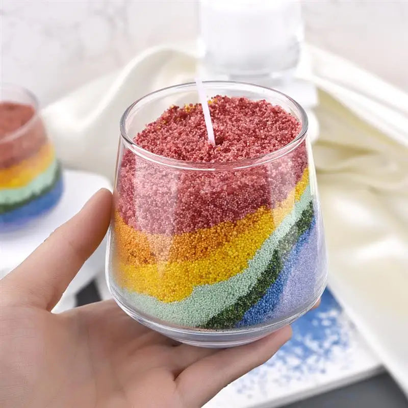 A hand holds a glass jar filled with layered colored sand pigment for a handmade candle wax kit.