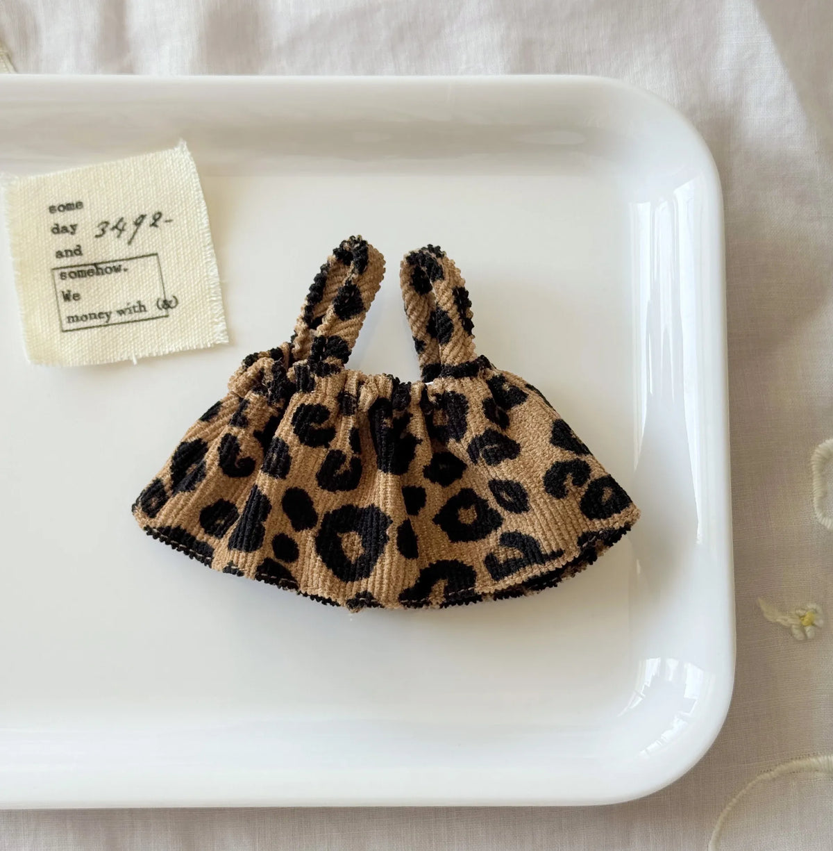 A leopard print shoulder strap skirt for dolls is displayed on a white plate next to a note.