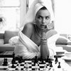 A woman in a white robe poses thoughtfully while playing a chessboard on a table.